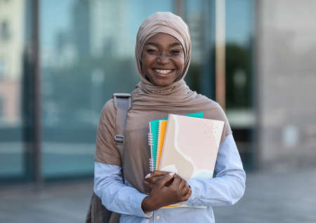 Education For Muslim Women. Smiling african islamic female college student posing outdoor with workbooks in hands, wearing hijab and carrying backpack, resting at campus, looking at camera, copy spaceの写真素材