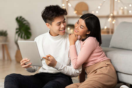Asian Millennial Couple Using Digital Tablet Computer Websurfing Online Sitting Together On Floor At...