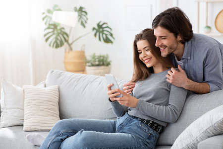 Positive young couple using mobile phone at home, happy woman showing her excited man creative ideas...