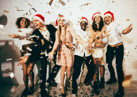 Christmas Party. Happy Young Men And Women Celebrating Xmas Wearing Santa Hats Standing Under Falling Confetti Having Fun Together Indoorの写真素材