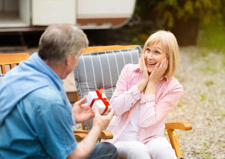 Senior man giving birthday gift to his beautiful surprised wife near RV in counryside
