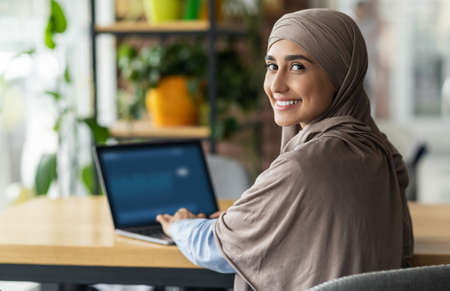 Back view of smiling muslim lady in hijab working or studying online at cafe, using laptop, copy space. Happy arab woman in headscarf freelancer typing on laptop, enjoying her distant jobの写真素材