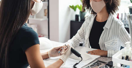 Regular client after opening salon after lockdown and procedures. Asian woman master in rubber gloves doing manicure with electric nail device for african american woman in modern studio, panoramaの写真素材