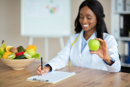 Cheerful African American nutrition adviser planning meals for client at clinic, copy space. Black female weight management specialist writing prescription for patient at hospital. Selective focusの写真素材