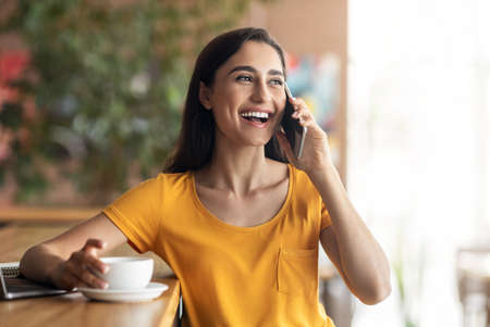 Happy young lady talking on mobile phone while drinking coffee at cafe, copy space. Cheerful arab woman having conversation on smartphone with girlfriend while spending time alone at cafeteriaの写真素材
