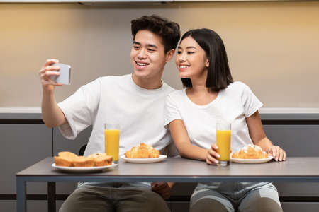 Happy Korean Husband Making Selfie With Wife During Breakfast Sitting In Kitchen At Home. Asian Family Couple Bonding Drinking Orange Juice And Eating Pastry On Weekend Morning Conceptの写真素材