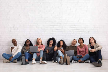 International group of friends with smartphones, laptops and digital tablets looking up on copy space, sitting on floor over white wall. Cheerful multiracial students using gadgets, studio backgroundの写真素材
