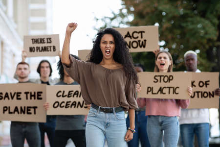 Save the nature. Mixed race young woman raising fist up and yelling, leading group of demonstrators on the street. Multiracial group of young people with placards protesting for healthy environmentの写真素材