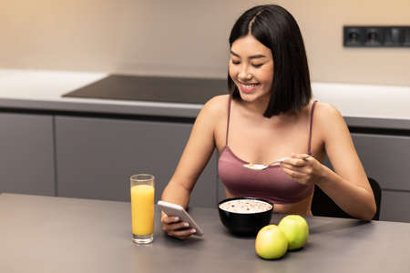 Weight Loss App. Fit Asian Lady Using Smartphone Eating Oatmeal Porridge For Breakfast Sitting At Table In Kitchen At Home. Healthy Diet, Nutrition For Slimming Concept. Empty Spaceの写真素材