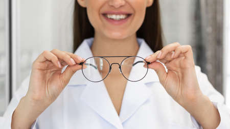 Eyesight And Vision Concept. Closeup of unrecognizable happy woman doctor showing new eyeglasses at camera, standing at optics store, blurred background. Female specialist optician offering specsの写真素材