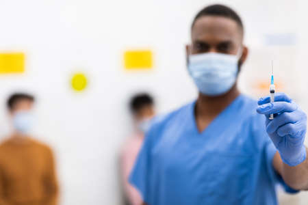 Covid-19 Vaccination. African American Doctor Holding Syringe With Coronavirus Vaccine Standing In Hospital, Wearing Protective Uniform And Mask. Blurred Background With Copy Space, Selective Focusの写真素材