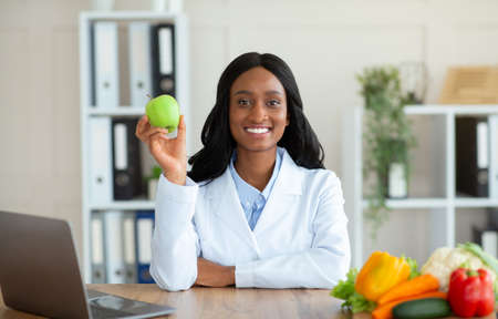 Positive black female dietitian holding apple fruit and looking at camera in clinic. Happy African American nutritionist with laptop offering online weight loss consultations at medical officeの写真素材