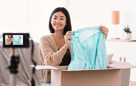 Fashion And Style Blog. Asian young woman vlogger recording unpacking video on camera device at home, female model influencer shooting brand advertising promo. Lady showing clothing after unboxingの写真素材