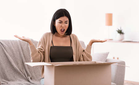 Bad Delivery Service Concept. Angry mad asian woman screaming and spreading hands after unpacking cardboard box, received damaged item, sitting on the couch, feeling upset with wrong parcelの写真素材