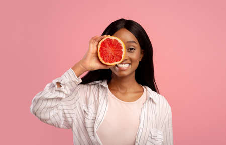 Vitamins for skincare and beauty. Pretty African American woman covering eye with grapefruit half on pink studio background. Black lady holding piece of fruit near her faceの写真素材
