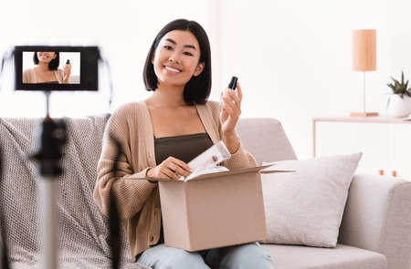 Lifestyle And Beauty Blog. Asian young woman vlogger showing cosmetics products, recording video on camera device at home, female model influencer shooting brand advertising promo. Unpacking, unboxingの写真素材