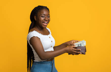 Finance, investment and money saving. Happy young black woman putting banknotes into wallet, planning budget, looking and smiling at camera, posing over yellow studio background with copy spaceの写真素材