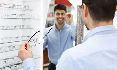 In Optics Market. Over the shoulder view of happy young man wearing glasses, looking in the mirror...