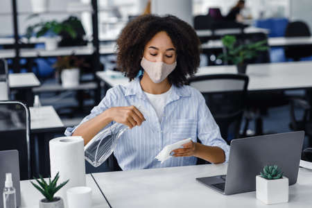 Workplace hygiene to protect health against coronavirus and new normal. African american woman in mask use spray and paper towels to disinfect desk and laptop in office interior, free spaceの写真素材