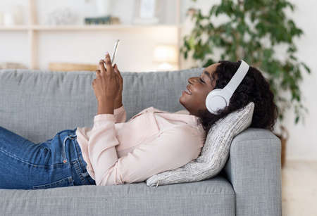 Relaxed Weekend. Happy Black Woman In Wireless Headphones Listening Music Or Browsing Internet On Smartphone While Resting On Comfortable Couch At Home, Lying On Sofa, Side View With Free Spaceの写真素材