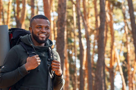 Smiling young african man with backpack hiking by forest, looking at copy spaceの写真素材
