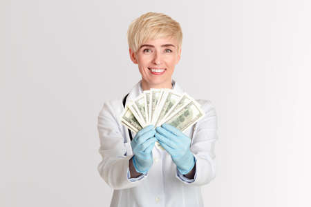Corruption of medical system and taking bribe. Happy female doctor in white coat and rubber gloves received money and shows dollars to camera, isolated on gray background, free space, studio shotの写真素材