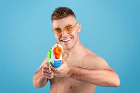 Summer vacation concept. Attractive millennial man pointing water gun at screen over blue studio background. Happy young guy having fun during his beach holiday on tropical resortの写真素材