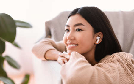 Enjoy The Music. Closeup portrait of calm asian woman wearing wireless earbuds listening to favorite song, relaxing on couch in living room. Smiling millennial lady lying on sofa and dreamingの写真素材