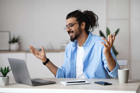 Excited indian man emotionally celebrating business success with laptop computer in office, sitting at desk and raising hands with joy, received good news, got promotion, finished project, free spaceの写真素材