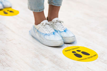Keep Safe Distance. Teen Girls Feet With Shoes Covers Standing Near Social Distancing Warning Sign On Floor In Queue Line Indoors. Covid-19, Coronavirus Protection Background. Croppedの写真素材