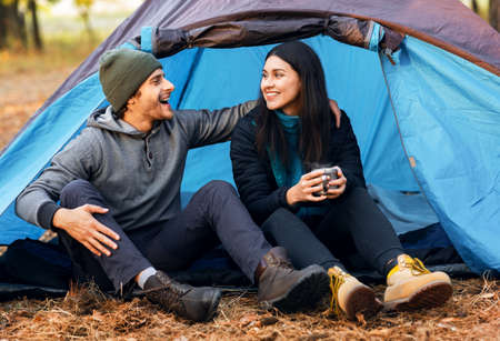 Romantic cheerful couple camping outdoors, sitting in tent, drinking tea and laughingの写真素材