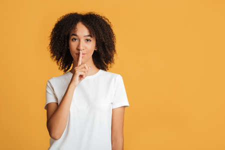 Millennial african american female in white t-shirt with serious expression shows hush sign, put finger on her lips isolated on orange background, studio shot, free space, cut outの写真素材