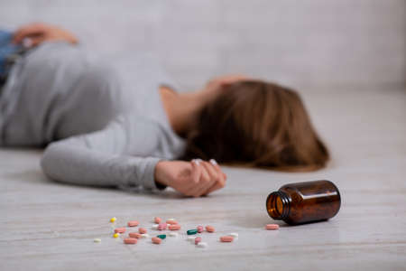 Young woman committing suicide by overdosing on pills, focus on bottle of tablets scattered on floor. Depression and drug overuse, mental breakdown or PTSD problem conceptの写真素材