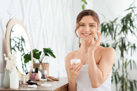 Happy young lady using face cream at home, looking at camera and smiling, copy space. Beautiful woman with ponytail holding cream jar and applying beauty product on her face, bedroom interiorの写真素材