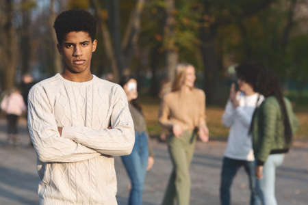 Depressed black guy with arms crossed standing apart from laughing multiracial students, public parkの写真素材