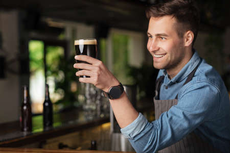 Professional work. Smiling bartender holds glass with dark craft beer and looks at it in interior of pubの写真素材