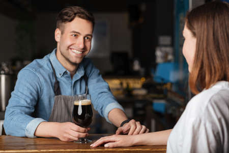 Communication with client in pub. Smiling handsome bartender gives glass of dark craft beer for girl in bar interiorの写真素材