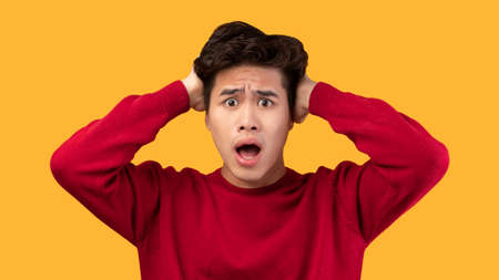 People, Human Emotions And Feelings Concept. Portrait of shocked scared asian man with open mouth grabbing head looking at camera in amazement, isolated over yellow studio backgroundの写真素材