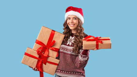 Happy young woman in Santa hat and Xmas sweater holding Christmas gifts on blue studio background,...
