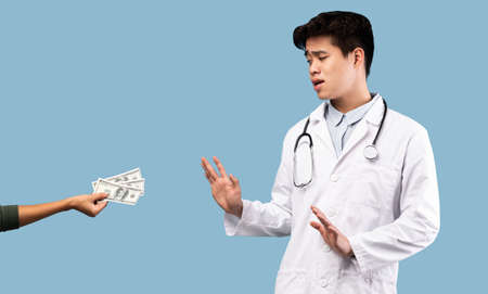 Serious scared asian male doctor in white coat refusing to take bribe from female patient giving him...