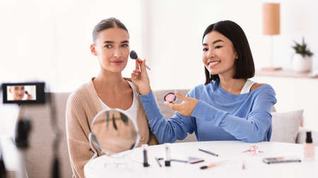 Young asian influencer applying blusher with brush on model, filming vlog for her internet channel in social media, looking at camera. Female make up artist putting cosmetic productの写真素材