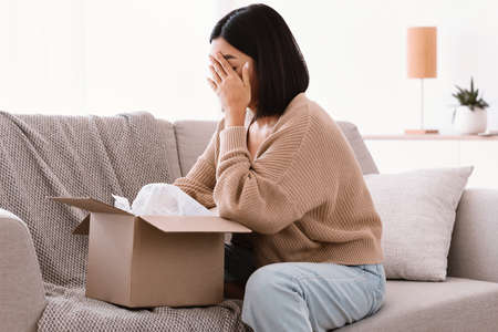 Side view of sad frustrated asian lady unpacking box, unhappy with bad delivery service sitting on...