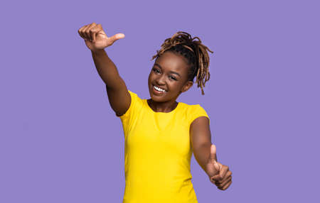 Joyful african american young woman smiling and showing thumbs up over purple studio background,...