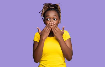 Happy emotional black woman covering her mouth with both palms and looking aside, purple studio background. Surprised african american young lady with eyes wide open looking at copy spaceの写真素材
