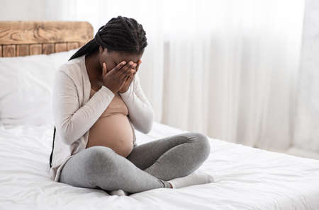 Hormonal changes during pregnancy. Black pregnant lady sitting on bed crying at home, feeling sad, upset expectant woman covering face with hands, suffering from prenatal depression, copy spaceの写真素材