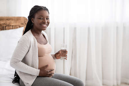 Happy African American Pregnant Woman Drinking Water From Glass While Relaxing On Bed At Home, Smiling Black Expectant Lady Enjoying Healthy Liquid, Thirsty Female Touching Belly And Looking At Cameraの写真素材