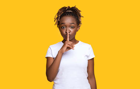 Pretty silent black woman making hush gesture, yellow studio background with copy space. Attractive african american young lady showing silence gesture, touching her lips with one fingerの写真素材