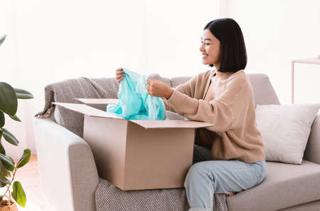 Happy Customer Concept. Excited asian lady opening and unpacking parcel cardboard box, holding blouse, sitting on sofa in living room at home. Young smiling woman satisfied with delivery serviceの写真素材