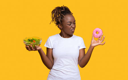 African american young woman looking at donut with disgust, holding fresh salad in another hand, yellow studio background. Healthy food, diet, nutrition concept. Good looking lady avoiding junk foodの写真素材