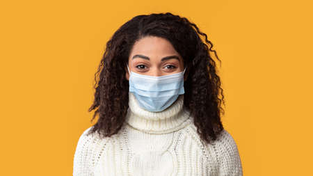 Infection And Protection Concept. Closeup front portrait of black young woman wearing medical mask isolated on pastel orange studio wall background. Glad smiling patient looking at camera, bannerの写真素材